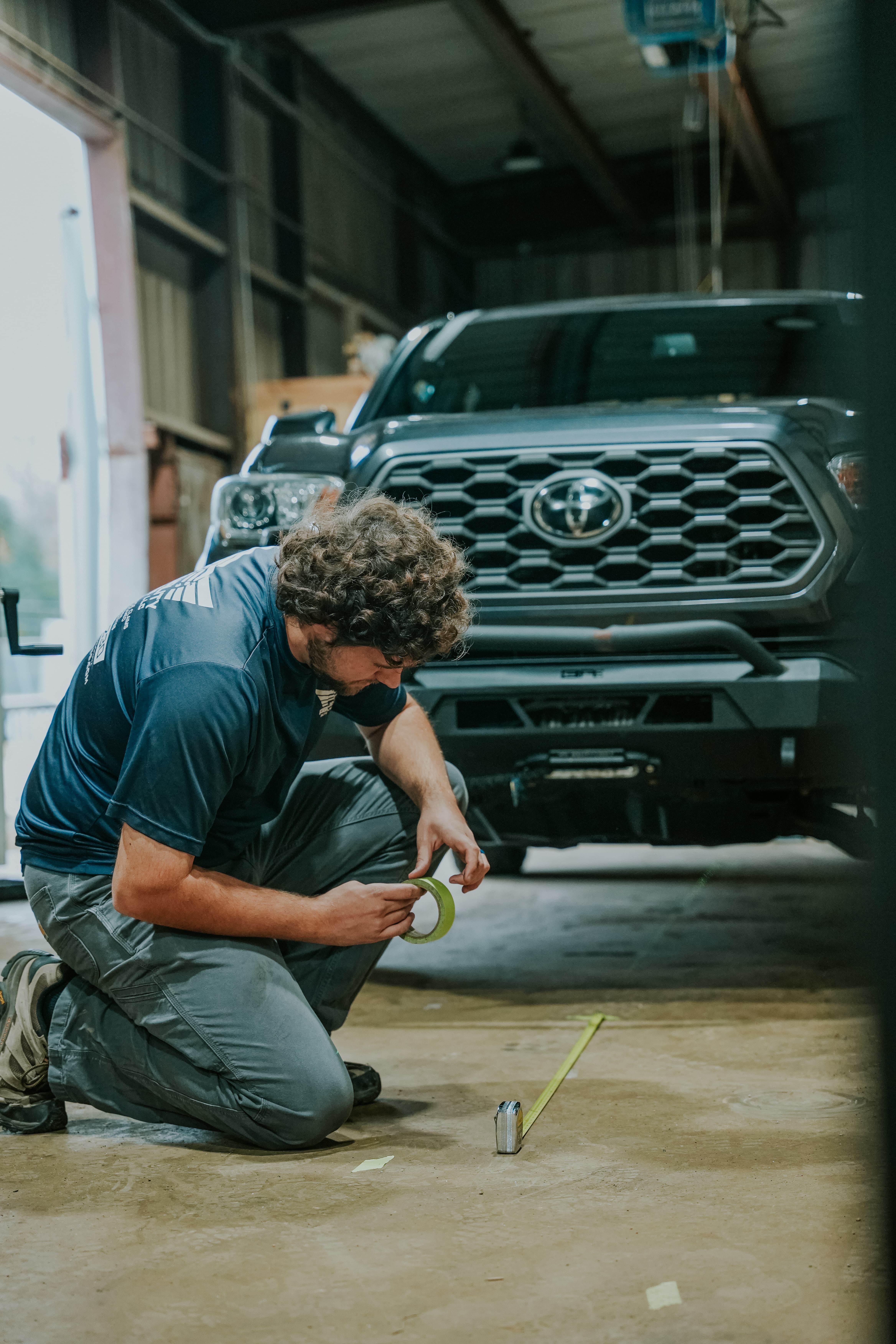 A mechanic kneels on a garage floor using a tape measure and masking tape in front of a dark gray pickup truck with a prominent grille, inside a well-lit automotive repair shop.