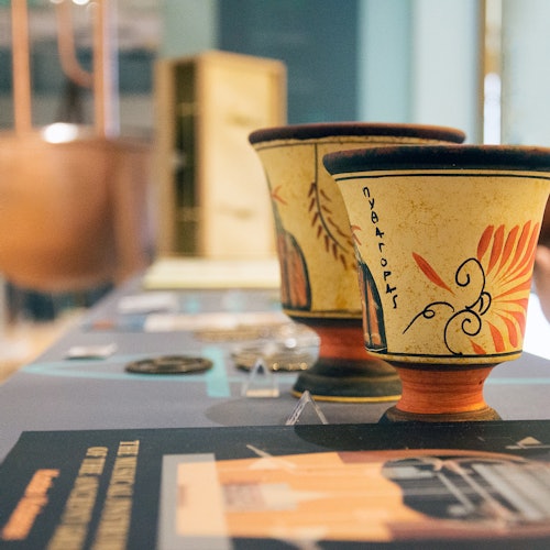 兩個飾有希臘風格圖案的陶瓷杯放置於展示桌上,四周擺放著各種小型文物。