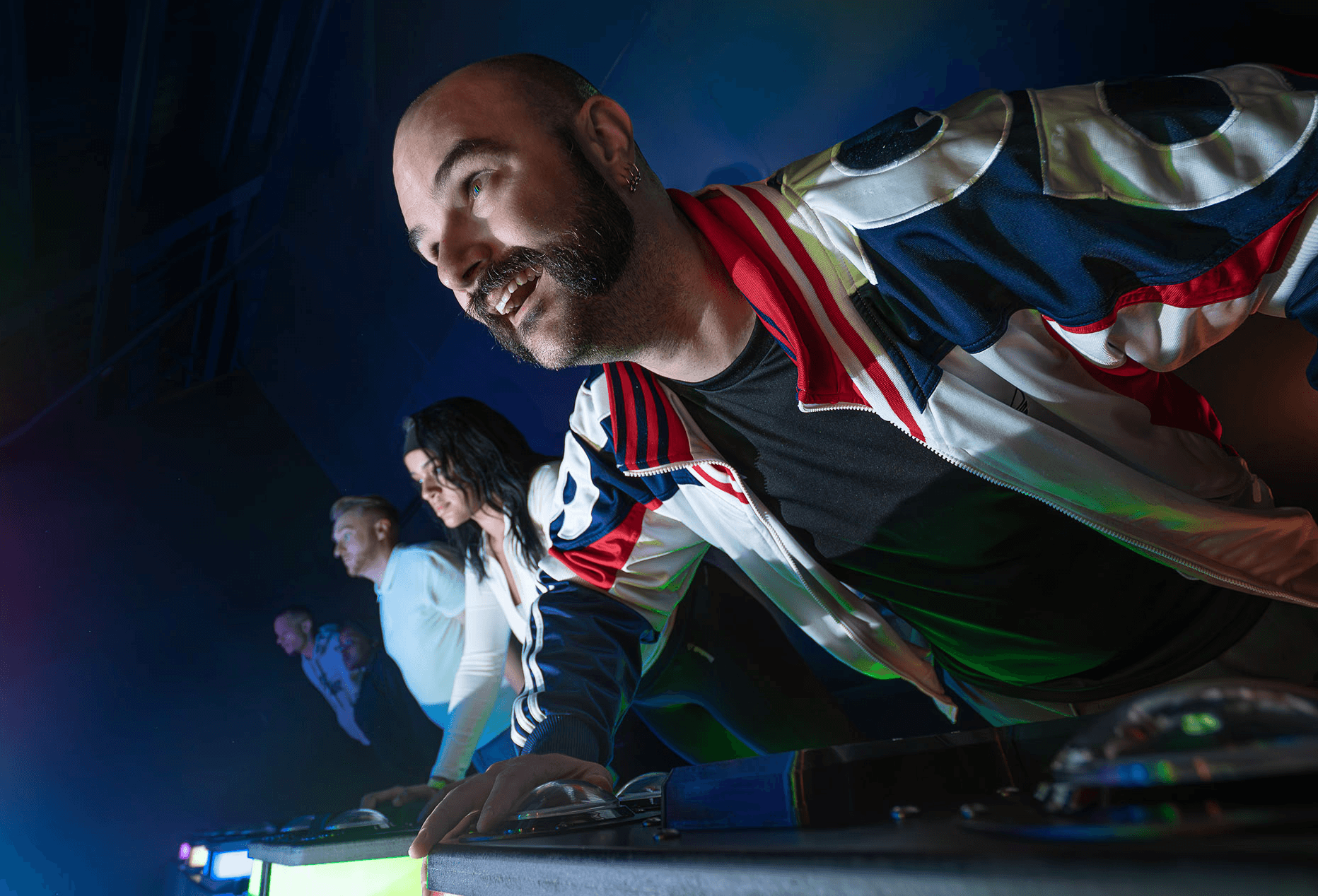 A low angle shot of a man with a beard leaning over a DJ mixer, other people are in the background.