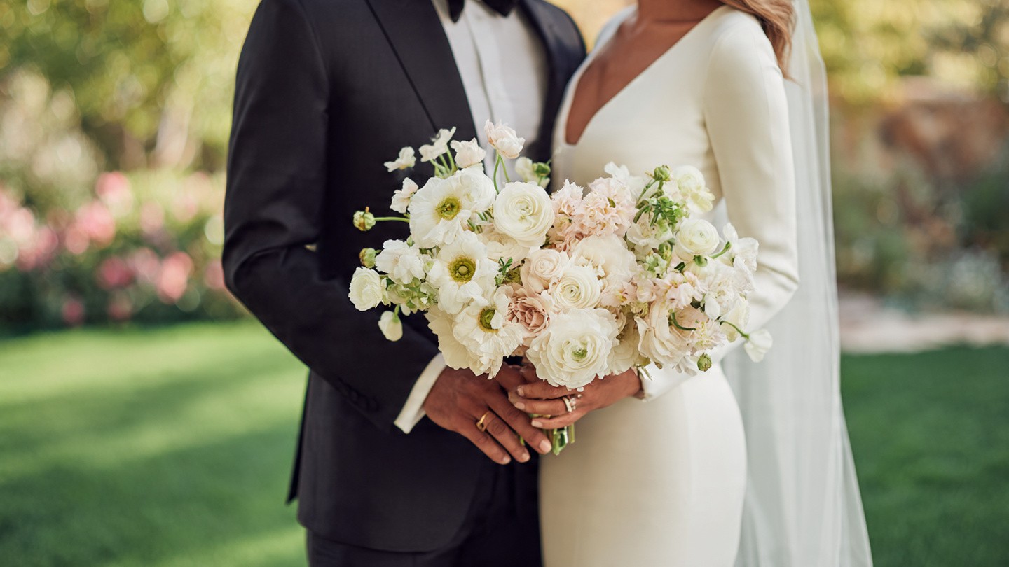 Warm golden-hour image of a bride and groom embracing; he wears a black tuxedo, she a deep V-neck long-sleeve cream wedding gown with veil; both hands clasp a luxurious oversized bouquet of blush, white, and champagne ranunculus with delicate greenery, set against a blooming garden backdrop.