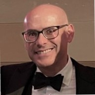 Professional headshot of a man with glasses and a dark blazer in a bright, blurred office setting.