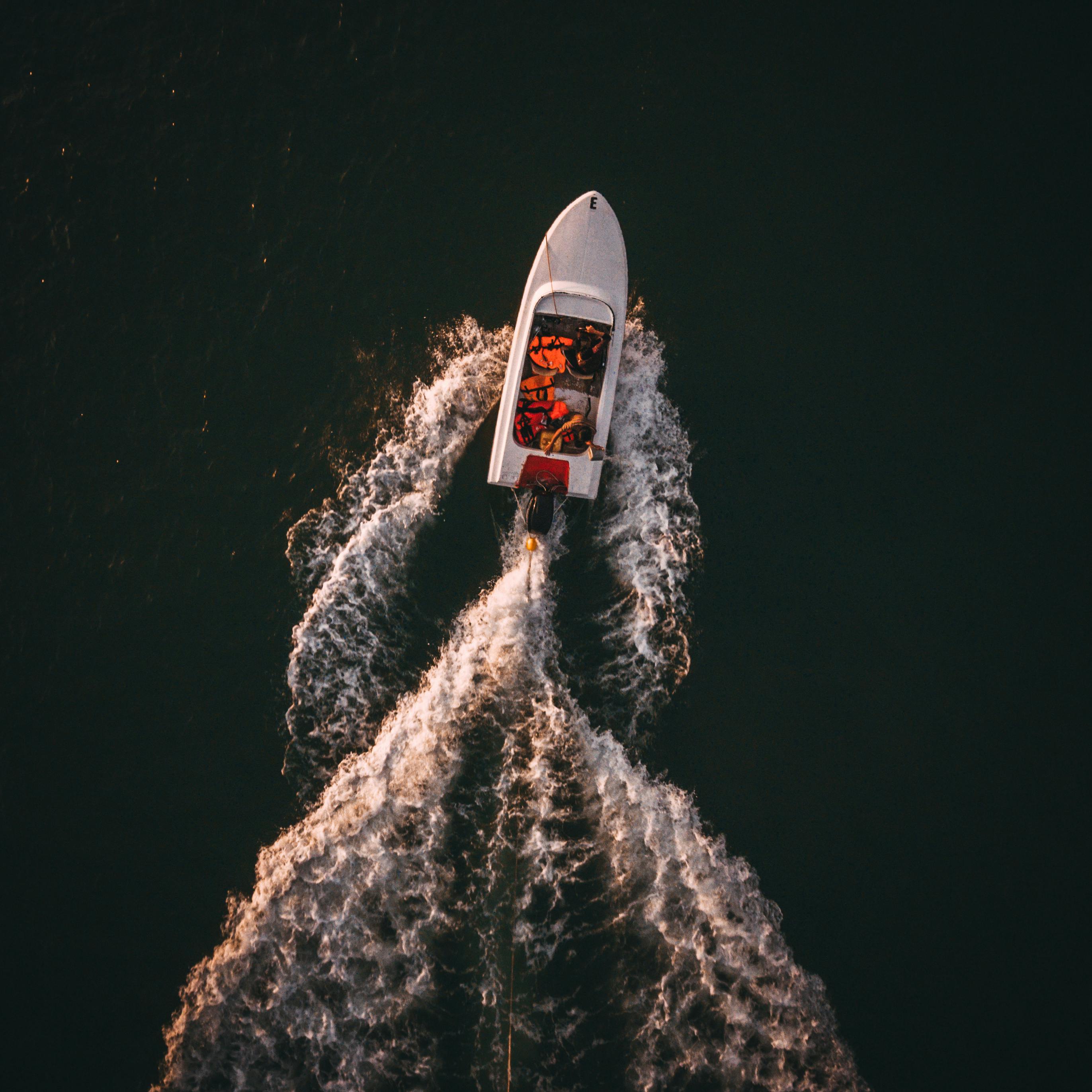 A white speedboat cuts through dark, rippling water, leaving a wake behind as it moves swiftly, with passengers wearing life jackets enjoying the sunny ride.