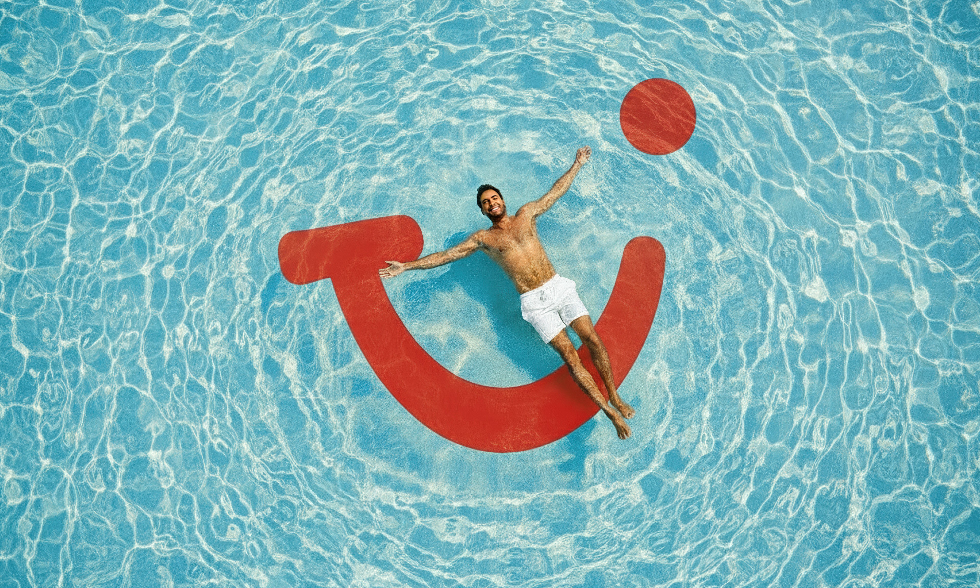 Eine Person liegt entspannt auf einer roten Luftmatratze in Form des TUI-Logos im hellblauen Wasser.