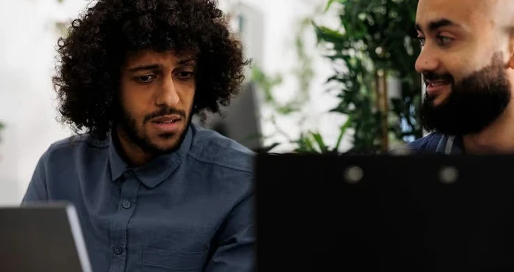 Dois homens lado a lado olhando para a tela de um laptop num ambiente escuro