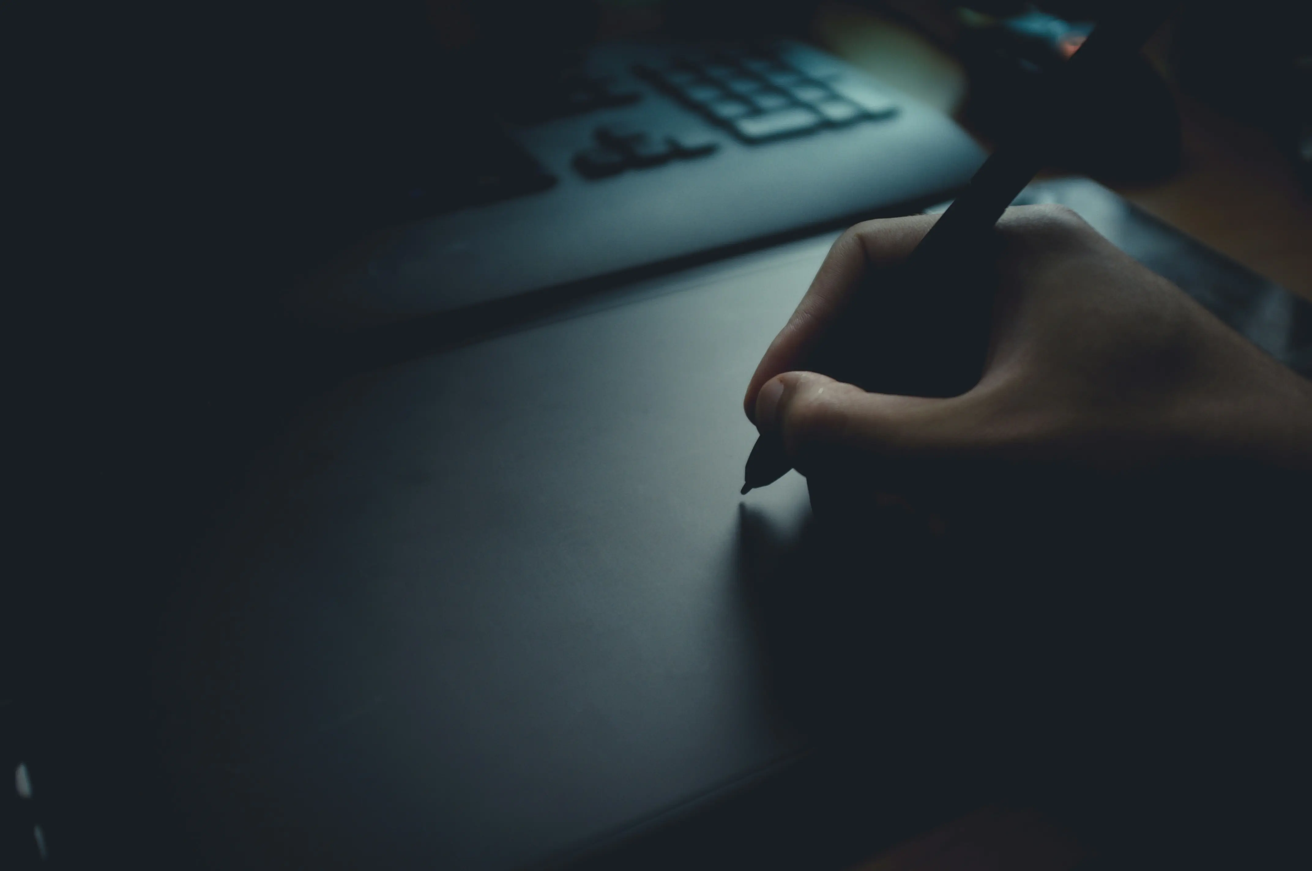 A hand holding a pen is seen above a dark desk, with electronic devices partially visible in the background.