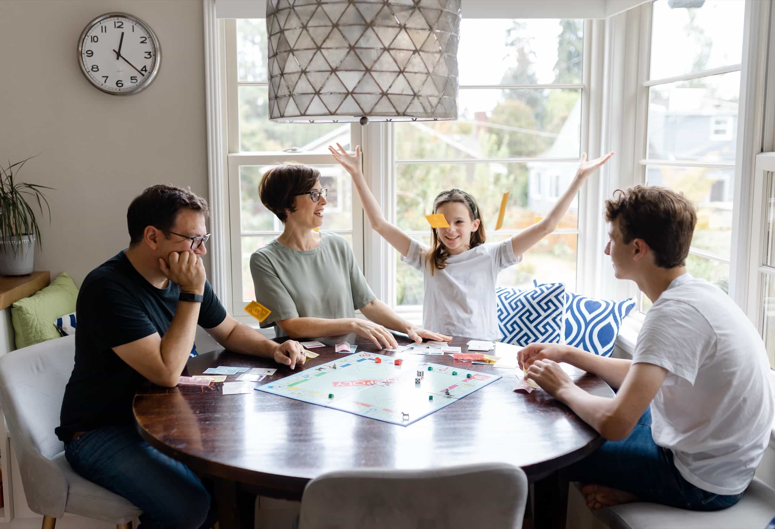 A family sits around a wooden table playing a board game in a bright, sunlit room, with a young girl joyfully raising her arms in the air.
