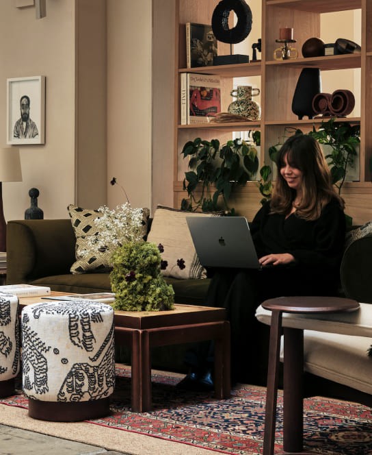 A cozy, well-decorated interior with a woman working on a laptop, surrounded by houseplants, eclectic furniture, and shelves filled with decorative items.