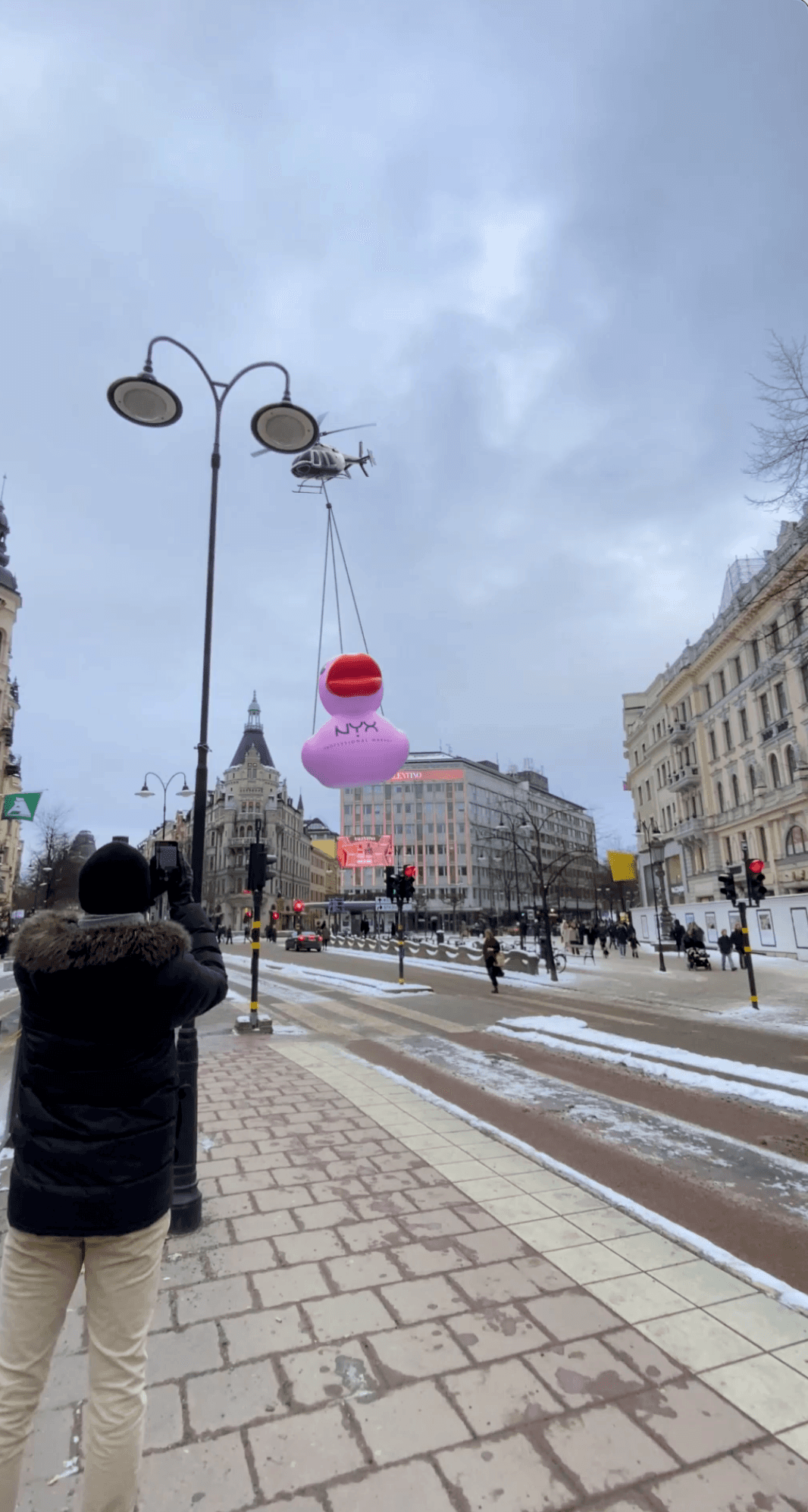 A helicopter carries a giant pink NYX duck with red lips over a snowy city street
