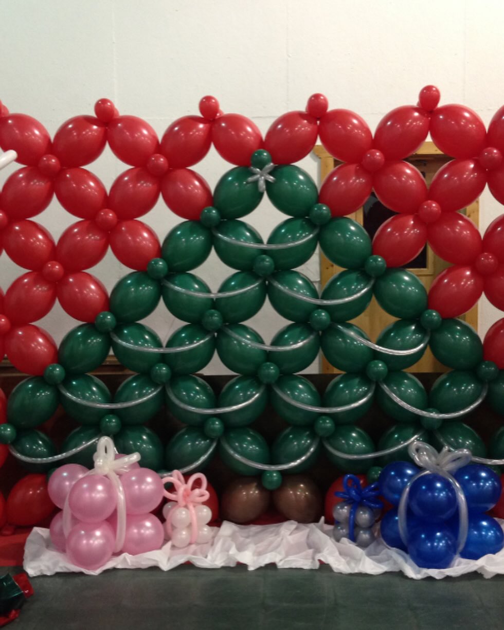 Red and green balloon wall with diamond pattern and gift boxes displayed at the base for a festive holiday setup by Be Bold Balloons