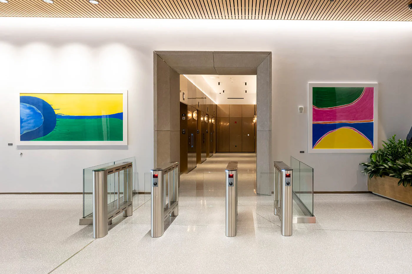 Office building lobby with polished terrazzo floors and security turnstiles, flanked by two bold abstract paintings — one featuring sweeping blue and green forms on yellow, the other a grid of vivid pink, green, blue, and yellow arcs.