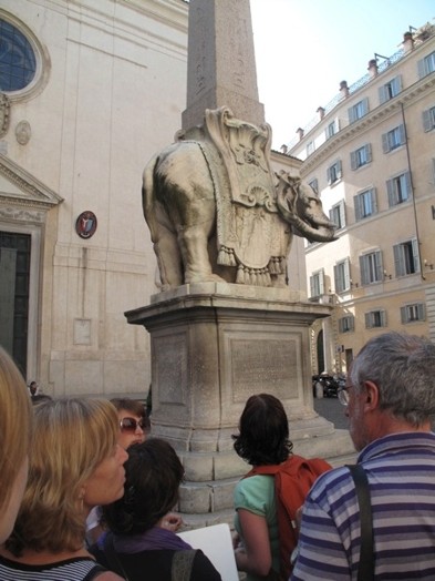 A group of people looking at a stone elephant that holds an obelisk on it.