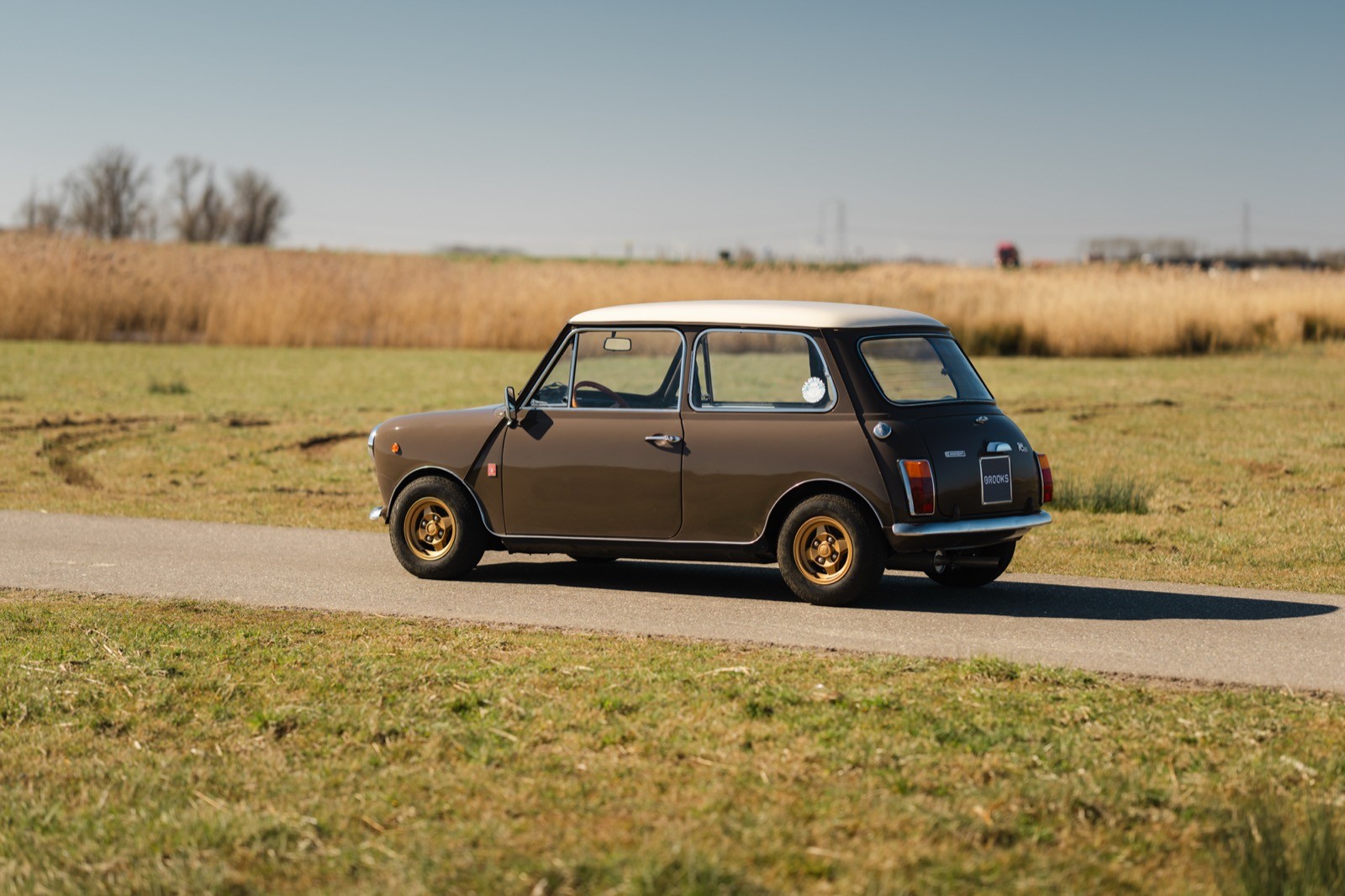 A vintage car parked on a gravel road, surrounded by open fields and a clear blue sky.