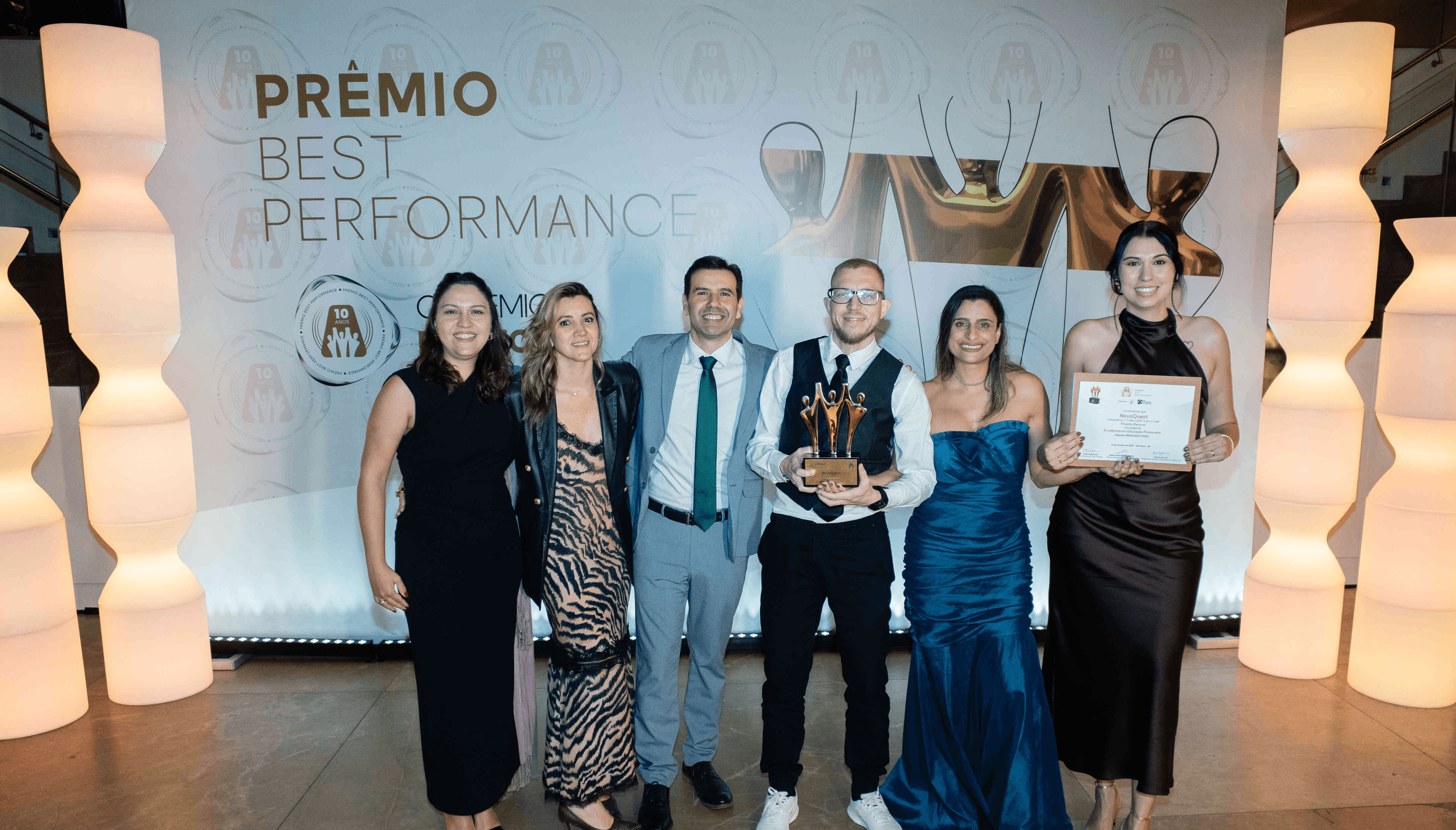 Executivos da NovaQuest posando com troféu ouro e certificado no palco do Prêmio Best Performance 2025