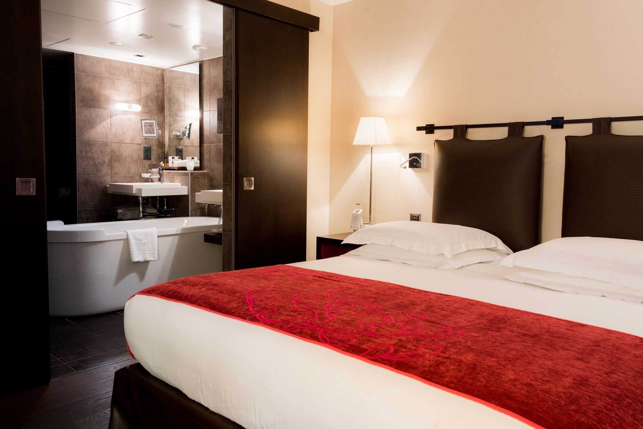 Bright hotel room featuring two neatly made beds with deep red throws against warm beige walls and modern bathroom fixtures.