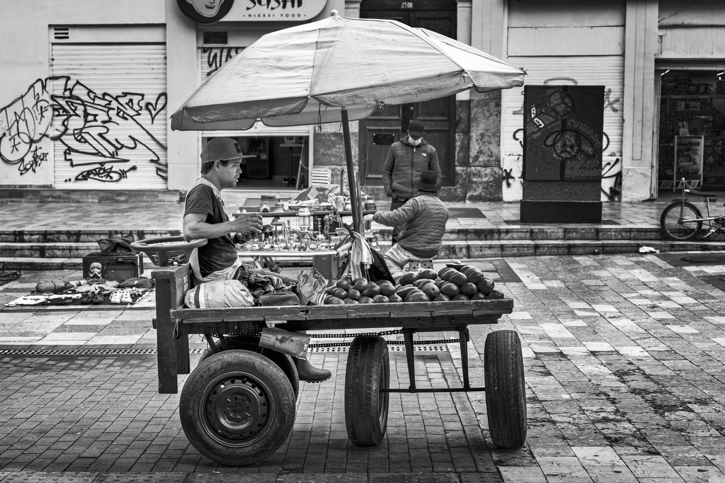 Black and white photo of a man seated behind a wooden fruit cart filled with avocados, shaded by a large umbrella, in a plaza surrounded by vendors and graffiti.