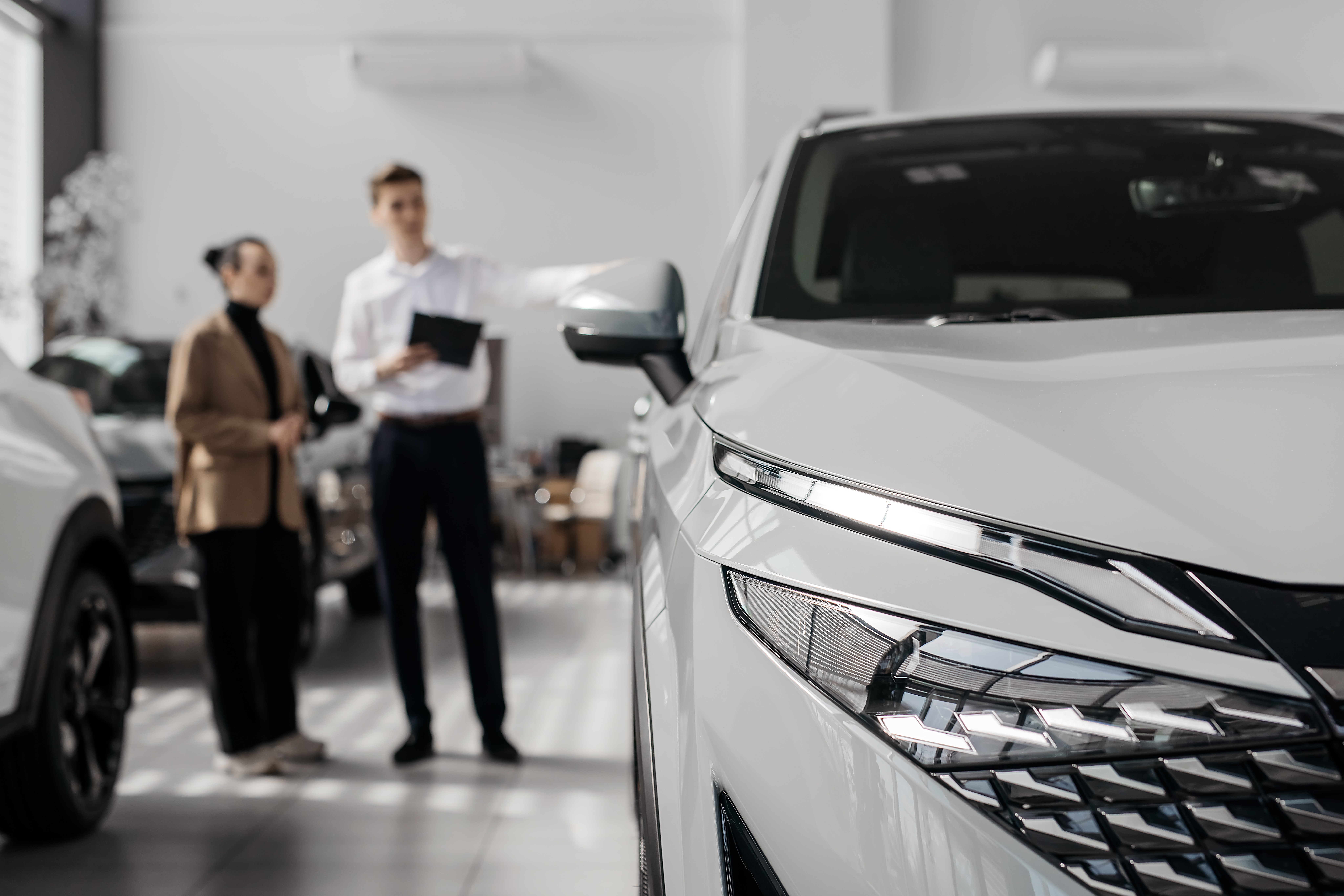 A car salesman in a dealership showroom with a female customer in the background and the front grille of a white sedan in focus in the foreground.