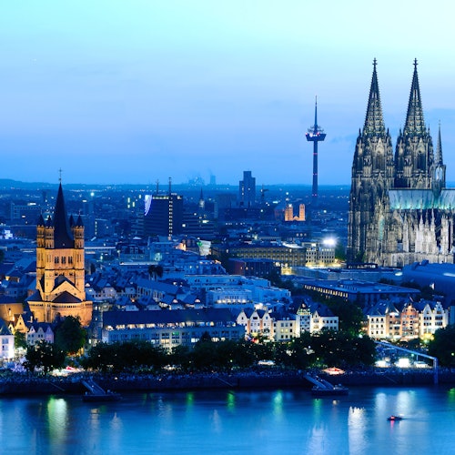 Pemandangan udara kota Cologne saat senja, menampilkan Katedral Cologne yang diterangi cahaya, Gereja St. Martin, dan Sungai Rhine di latar depan.