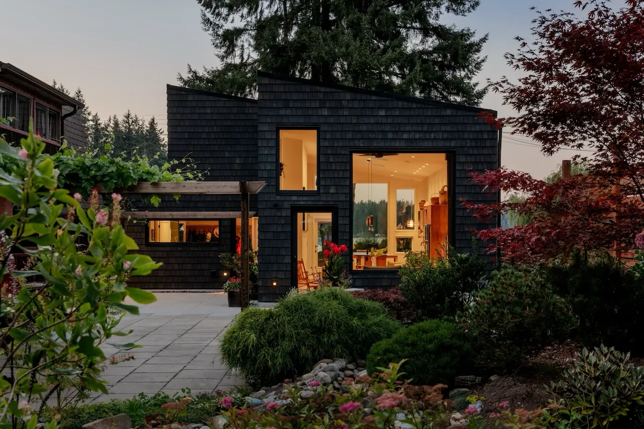 Modern two-story home with large glass windows, warmly lit interiors, and a balcony. Surrounded by trees and a manicured lawn at dusk. Cozy ambiance.
