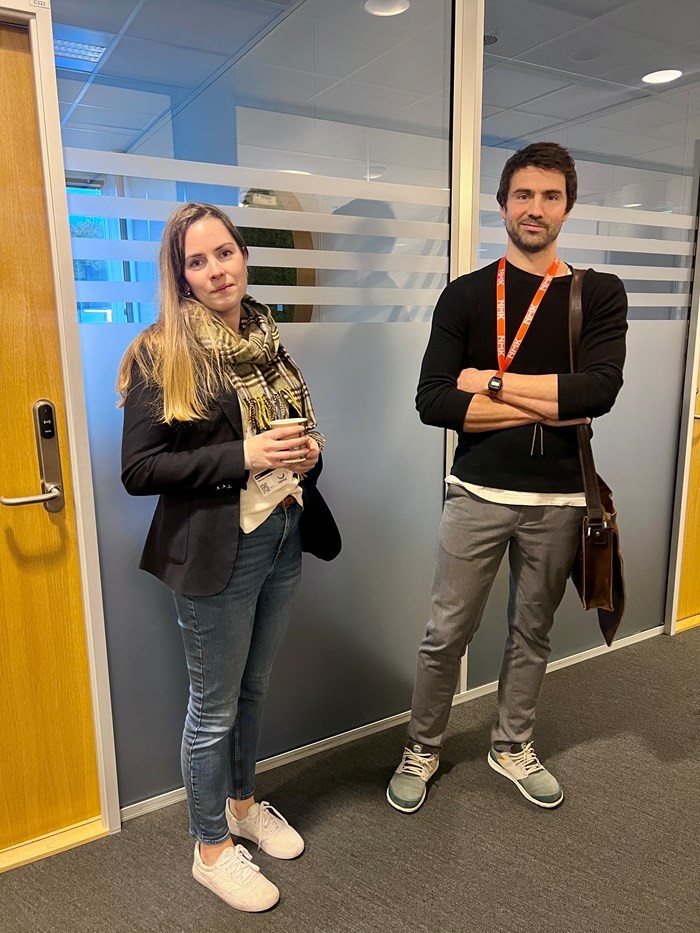 Two young adults stand smiling in front of a glass wall, each holding a coffee cup. Informal attire is worn.