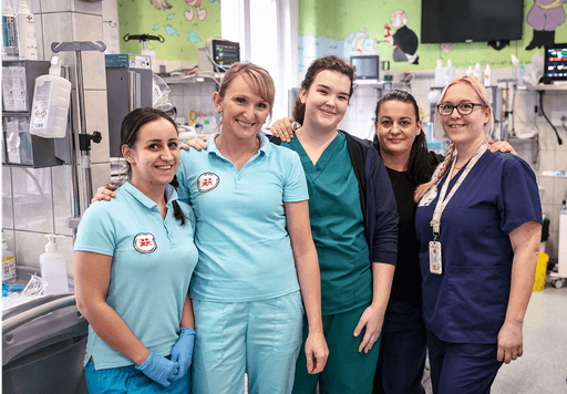 Mosolygó egészségügyi dolgozók csoportképe kórházi környezetben, színes gyerekmintás faldekorációval a háttérben.