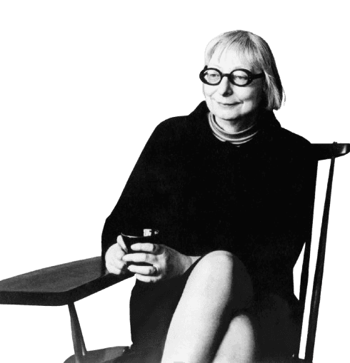 A black-and-white photograph of a woman with short, light hair and large, round glasses, seated in a modern chair with a relaxed, thoughtful expression. She is dressed in a dark outfit, holding a small cup, with abstract artwork visible in the background. Her attire, pose, and surroundings suggest a creative or intellectual persona, likely from the mid-20th century.