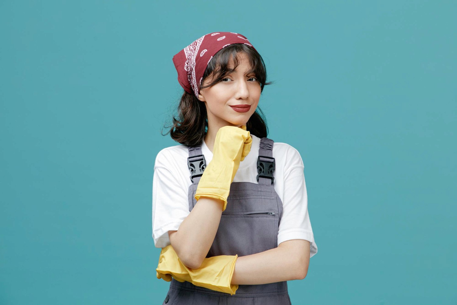 Confident female cleaner wearing yellow gloves and a red bandana, posing against a blue background, representing professional cleaning services.