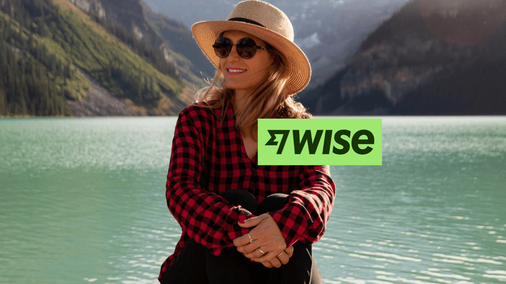 A professional photo of a newly landed newcomer to Canada, wearing a red plaid shirt and straw hat, seated by a stunning turquoise glacial lake in the mountains. A high-quality embroidered Wise logo patch (dark green text and icon) is subtly integrated onto her shirt, with a detailed physical Wise Visa card partially visible, representing managing travel finances.