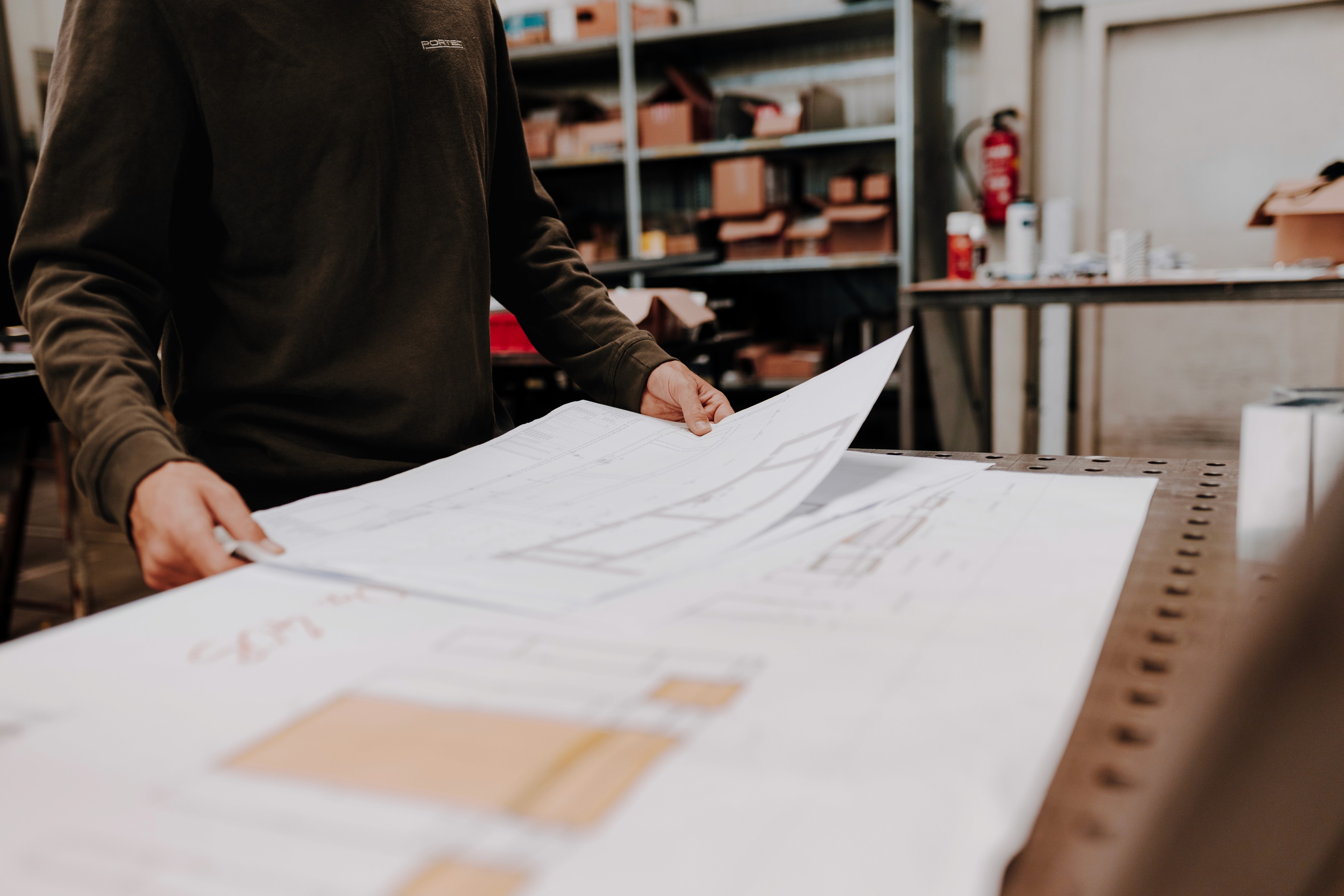 Person reviewing architectural plans, holding paper, in a workshop setting with a metal table.