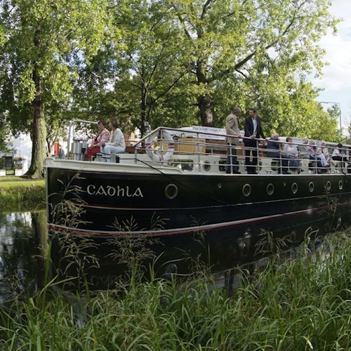 A boat named "Caolhla" carrying several people navigates a serene, tree-lined canal. Passengers are seated and standing on the deck.