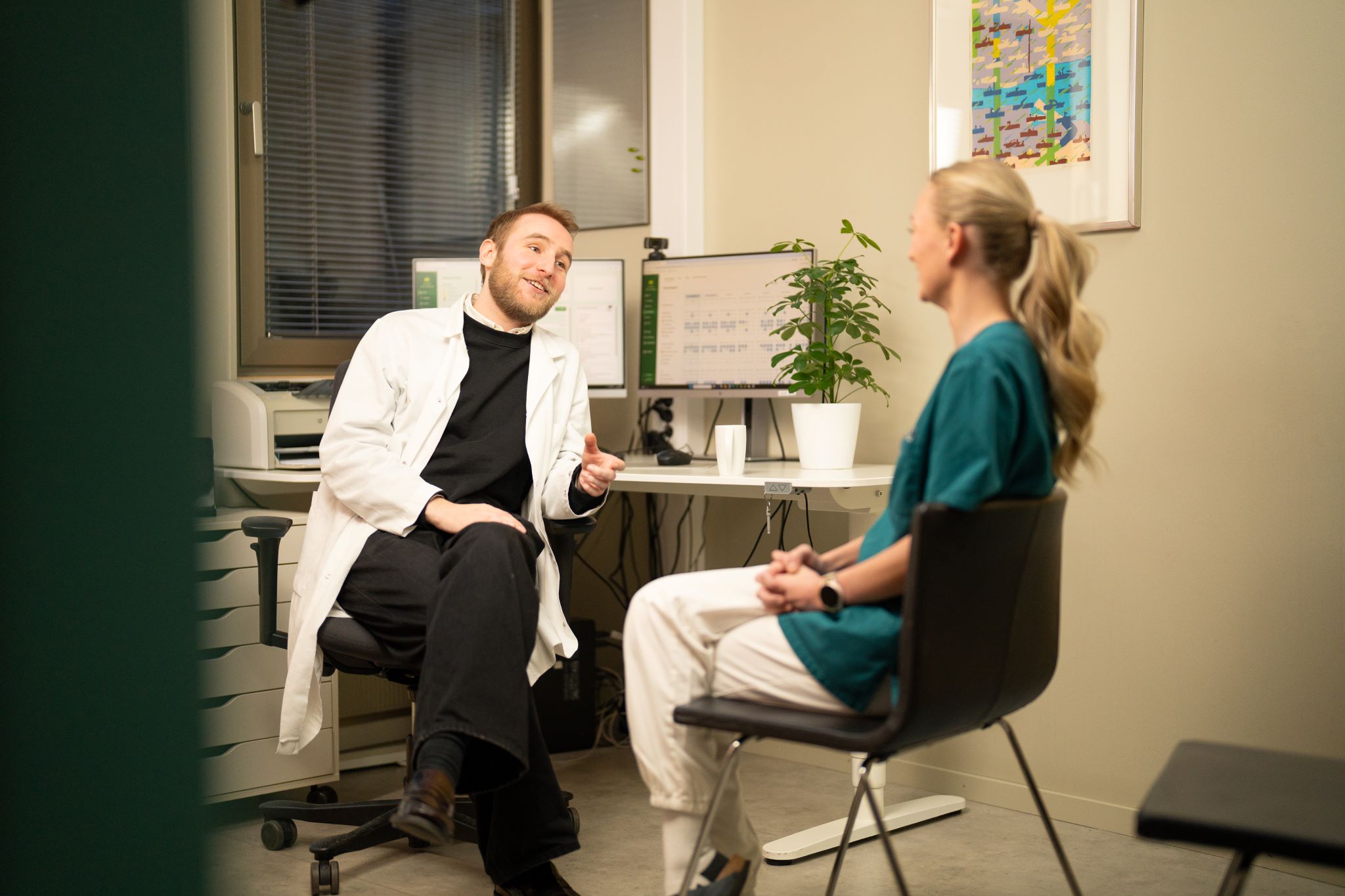 En lege i hvit frakk og en helsesekretær sitter overfor hverandre på et moderne kontor og snakker.