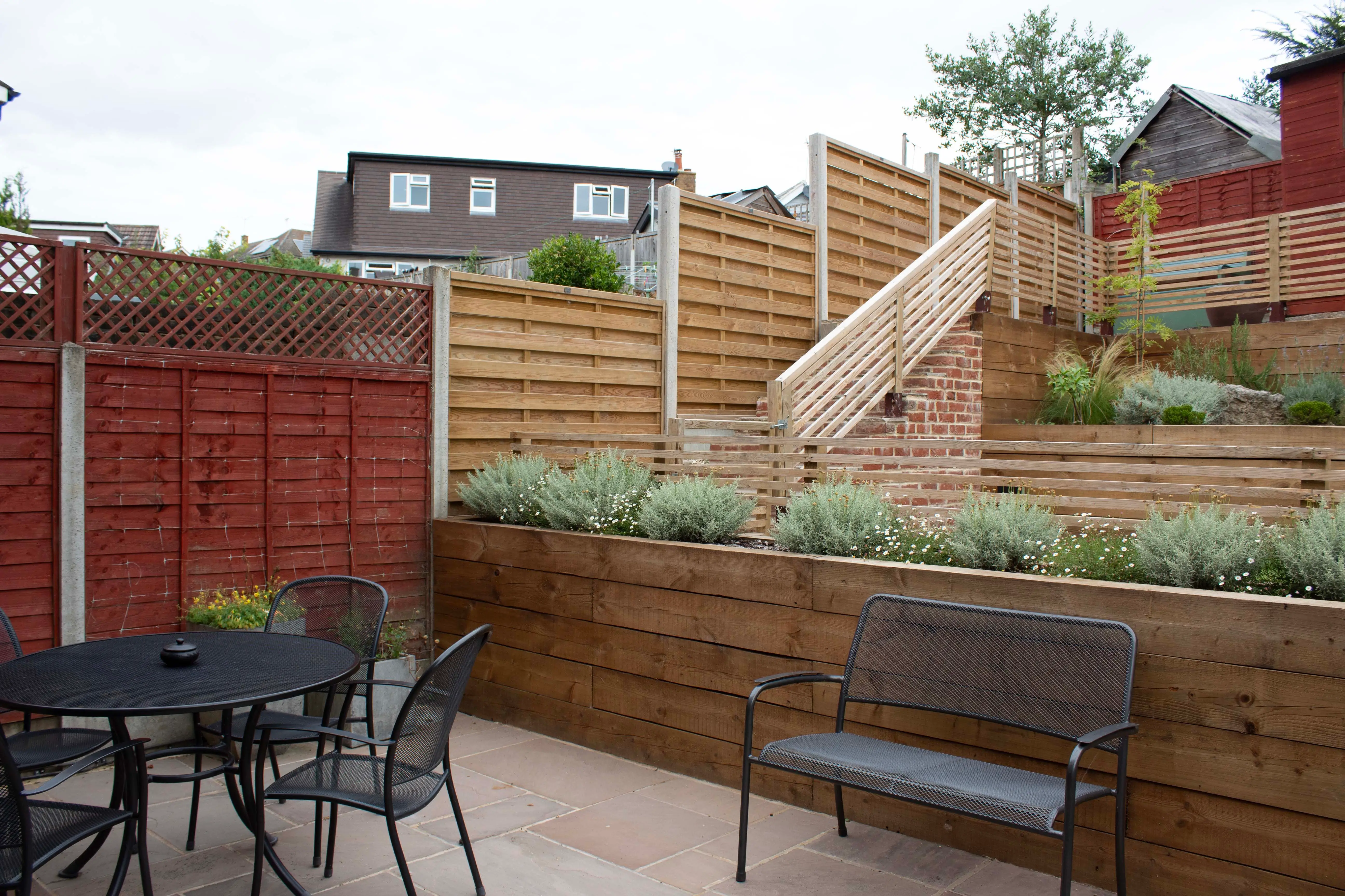 A backyard view featuring wooden fences, a patio with a table and chairs, and steps leading to a grassy area.