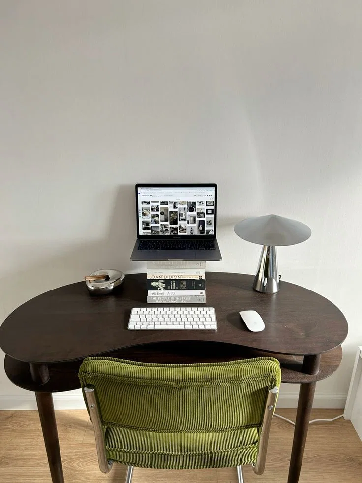 Un pequeño escritorio con un ordenador portátil, un cuaderno y una lámpara, con una silla verde contra una pared lisa.