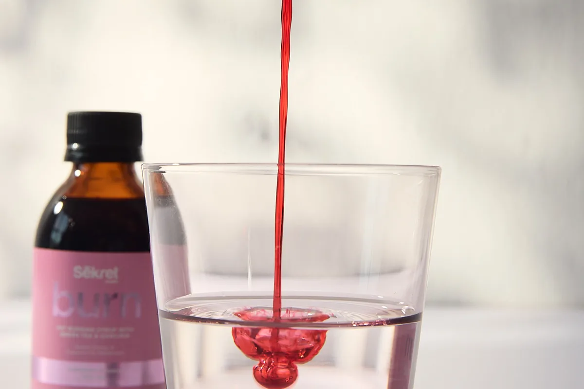 Red Sēkret Burn syrup poured into a glass of water beside its bottle
