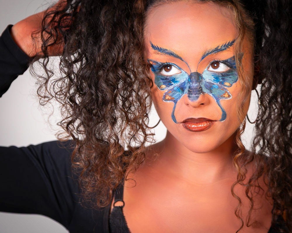 Close-up dramatic beauty portrait of a woman featuring detailed blue and orange butterfly face paint around her eyes, looking directly into the camera.