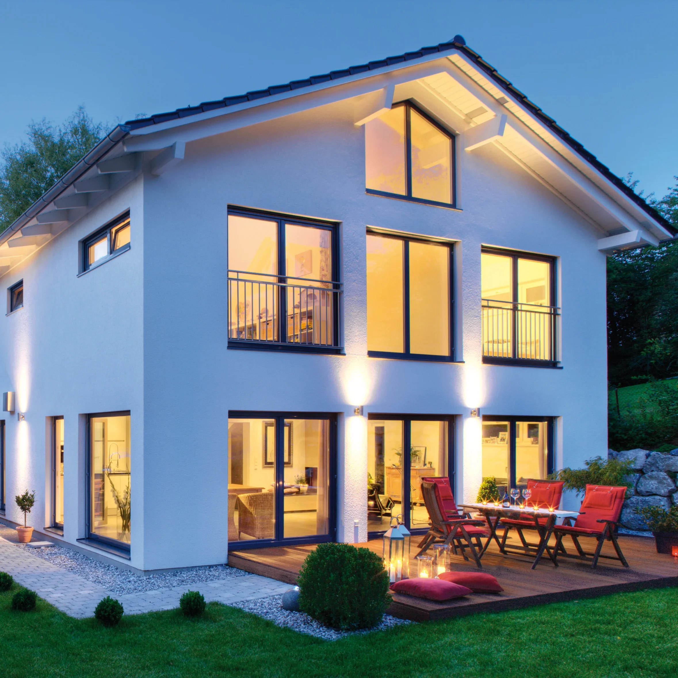 Ein modernes, beleuchtetes Einfamilienhaus mit weißer Fassade, großen Fenstern und einer angrenzenden Holzterrasse mit Sitzgelegenheiten.