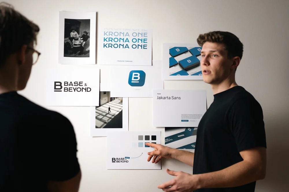 Felix Maier und Paul Schlager diskutieren vor einer Moodboard-Wand mit Markenlogos, Farbschemata und Typografie-Vorlagen von „Base & Beyond“ – professionelle Markenentwicklung und visuelle Identität.
