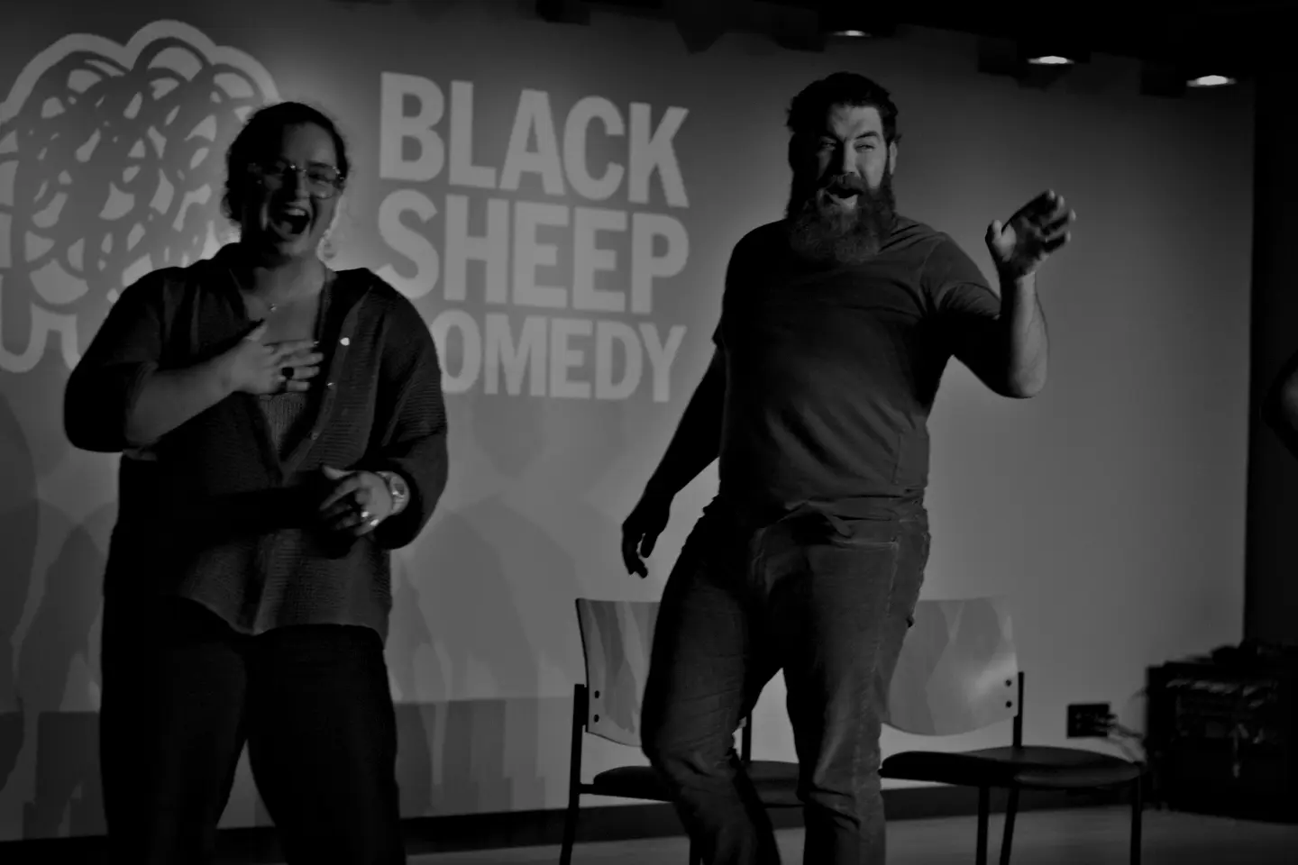 Two performers laughing and interacting with the audience on stage at Black Sheep Comedy, with the Black Sheep Comedy logo visible behind them. Capturing the fun and energy of live improv and sketch comedy performances in Carrboro and Chapel Hill.