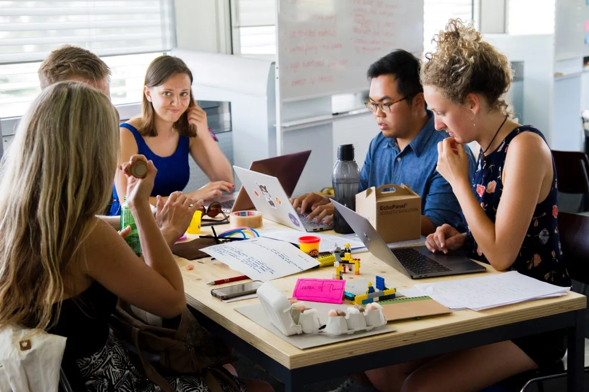 Group of five young adults collaborating at a workspace table, surrounded by laptops, notes, craft materials, LEGO pieces, and a whiteboard in the background, suggesting a creative team brainstorming or prototyping session.