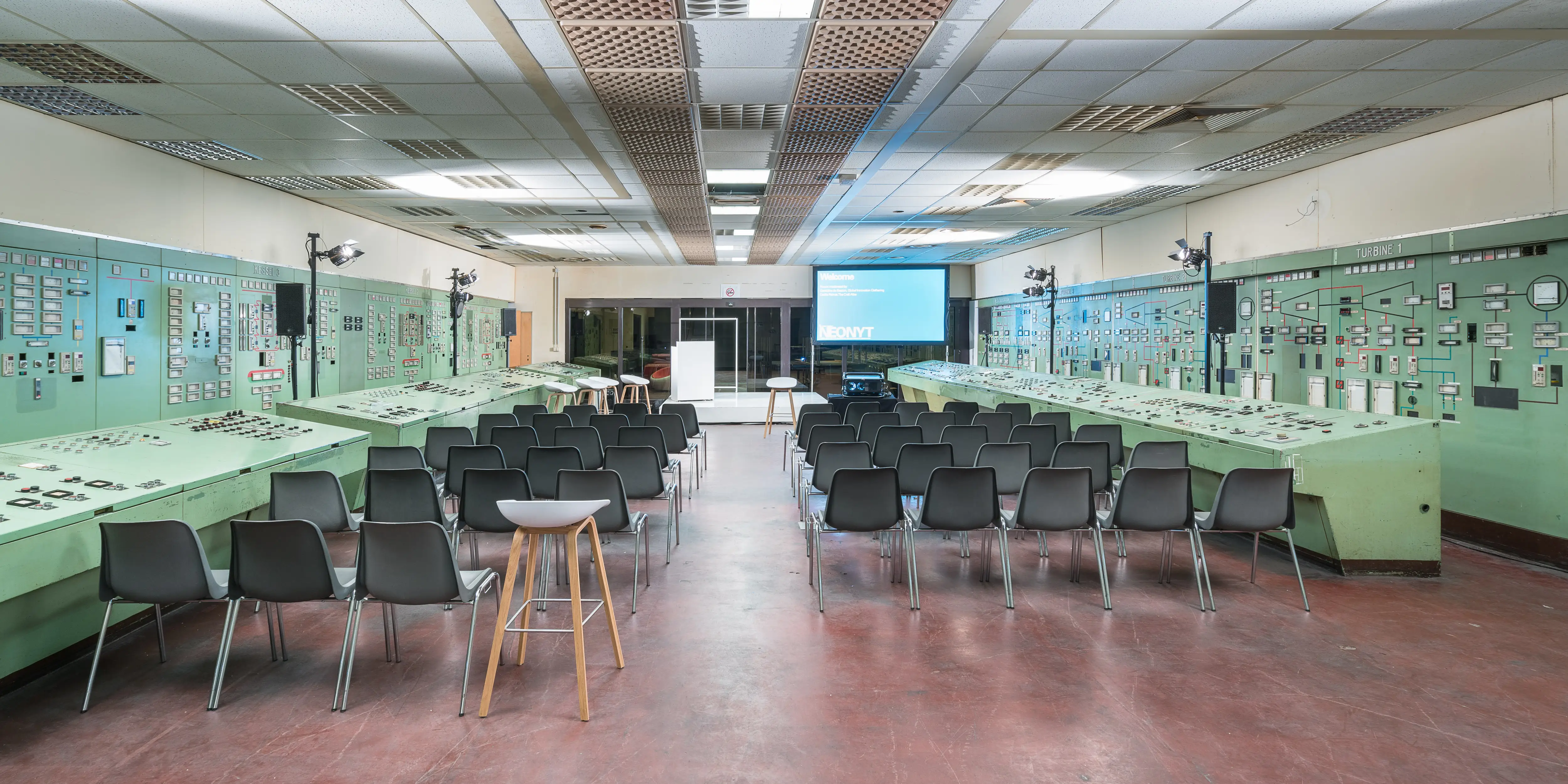 Workshop space in the historic control room with mint-green instrument panels, black chairs and a presentation screen at NEONYT.