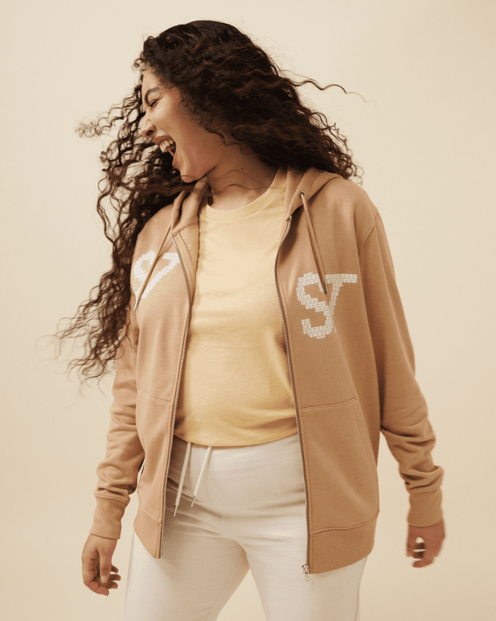 Woman with long curly hair laughing while wearing a tan zip-up hoodie with a logo.