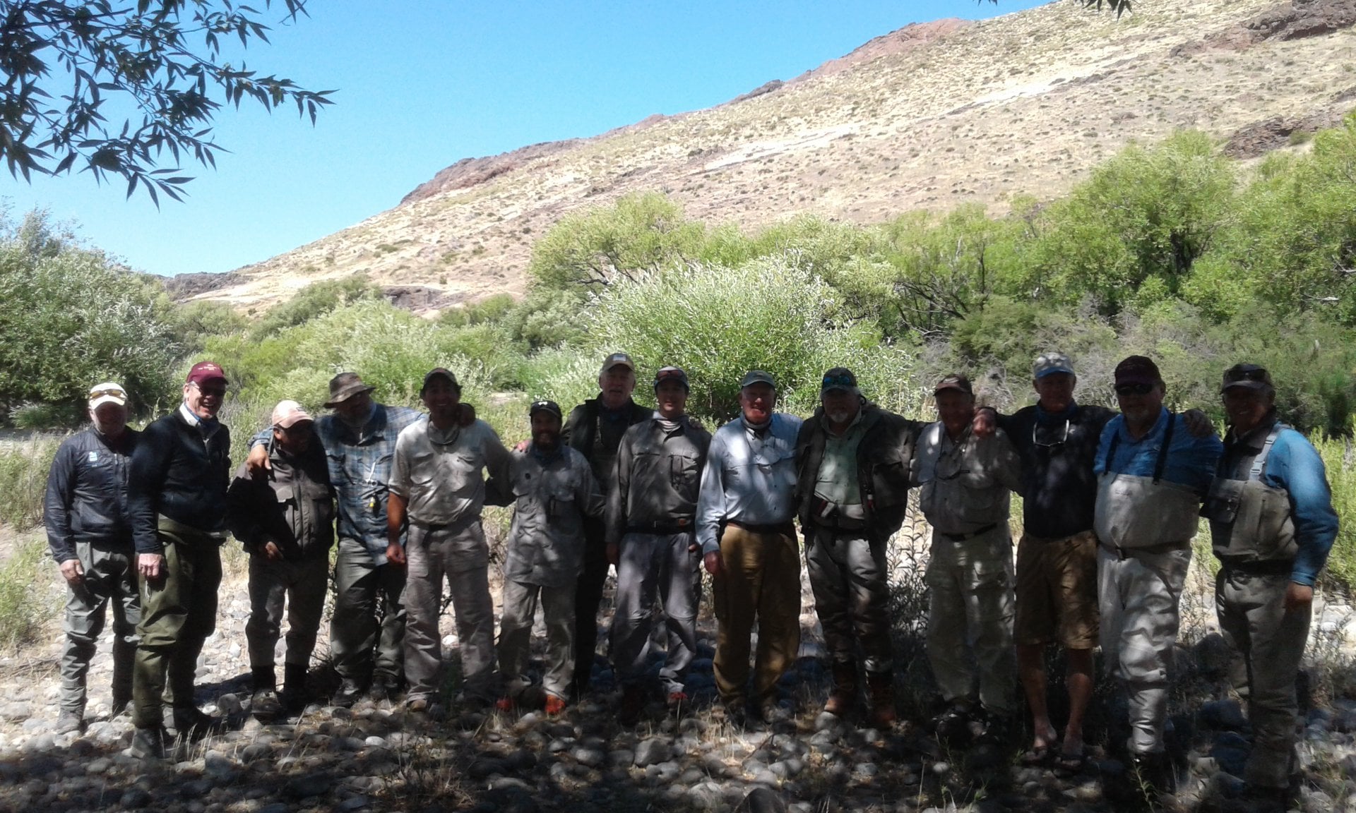Gran Grupo de pescadores, cerca de 20, en la orilla del río un día soleado, con las montañas de fondo