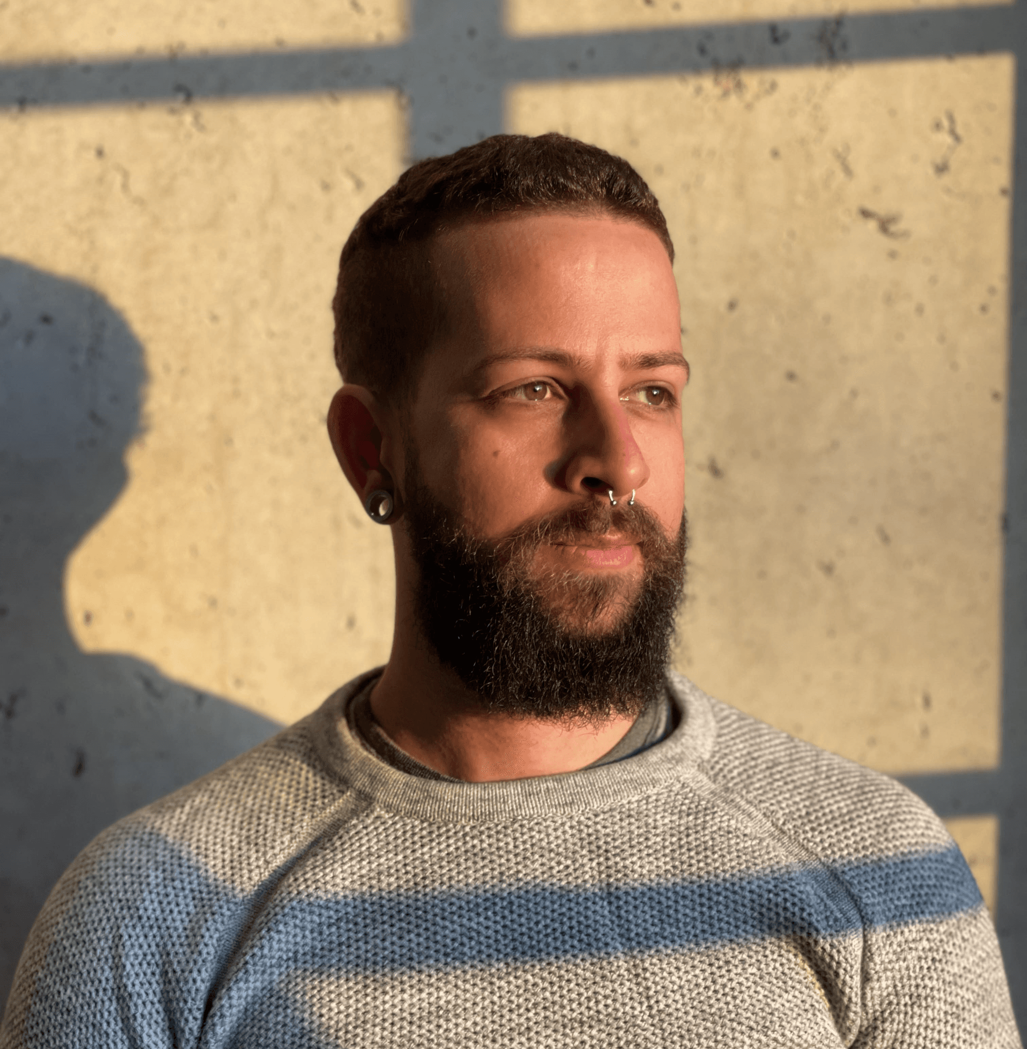 David: A bearded man with a septum piercing is illuminated by natural light, standing against a concrete wall. He wears a textured sweater and gazes thoughtfully to the right.