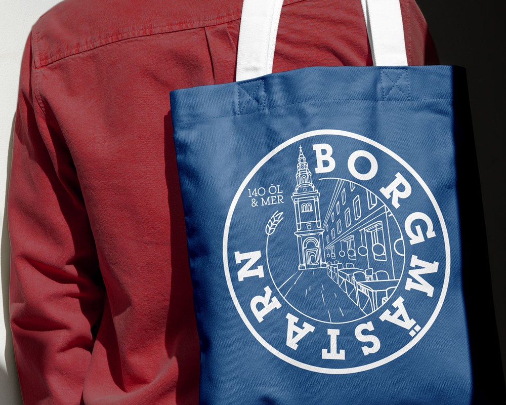 A person in a burgundy sweatshirt holding a navy blue tote bag with the white Borgmästarn circular logo, showing the brand identity applied to merchandise.
