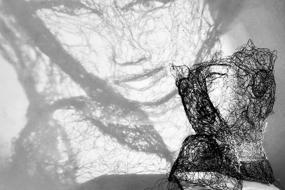 Sculpture en fil de fer d’un buste de femme, éclairée pour projeter un visage en ombre sur le mur