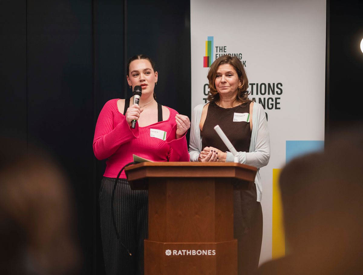 Person in blue trousers, mustard jacket, and white sneakers against a split orange and white wall.