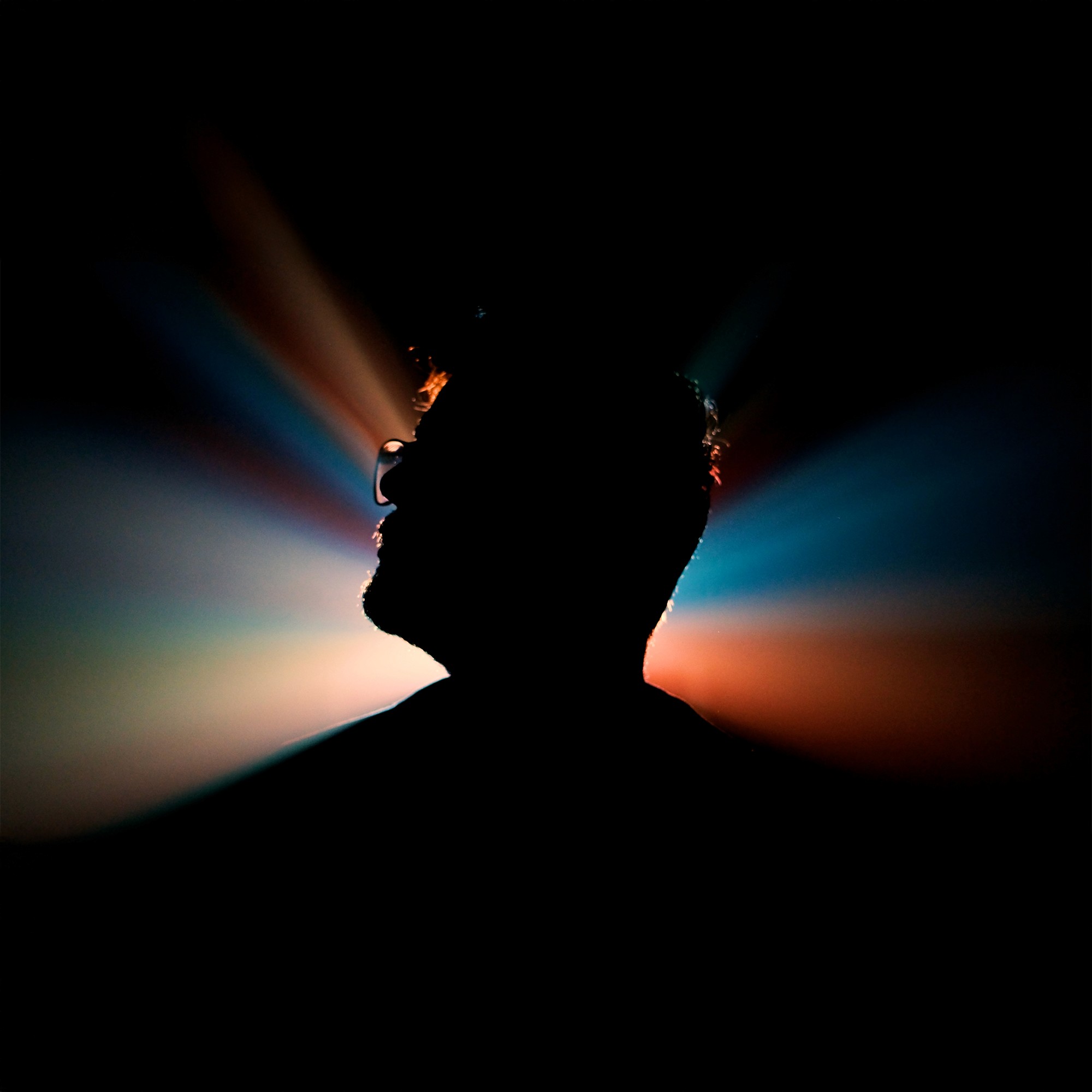 A young person with brown hair, wearing reflective silver sports sunglasses and a black sweater, looks at the camera while resting their chin on their hand.