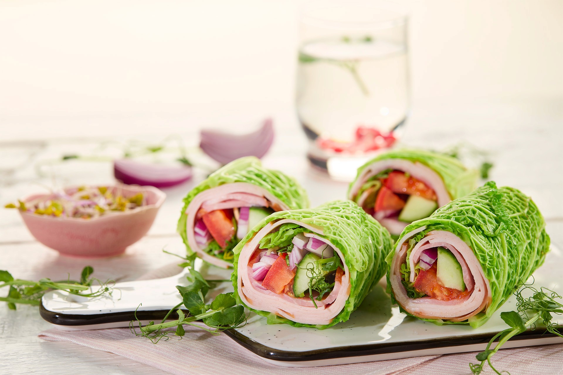Fresh lettuce wraps filled with turkey, cucumbers, tomatoes, and red onions are neatly arranged on a white serving platter with a bowl of sauce and a refreshing glass of infused water in the background.