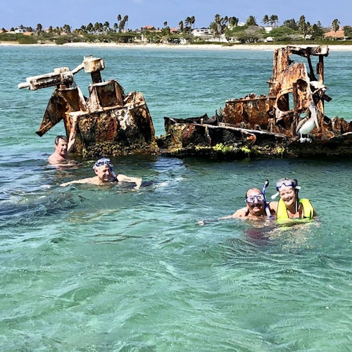 Quatre personnes font de la plongée avec tuba dans une eau claire et turquoise près d'une épave rouillée, avec un rivage lointain en arrière-plan.