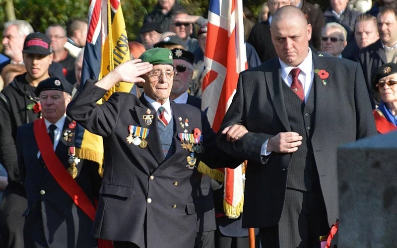 A photograph at a remembrance day event, many people in military dress in the background and in the foreground a man in a suit with short hair, wearing a poppy on his lapel stands arm in arm with an older man wearing a green beret and many medals, he is saluting
