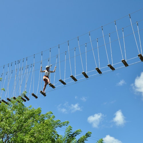 A person wearing safety gear navigates a suspended rope bridge high above the ground against a clear blue sky. Trees are visible below.